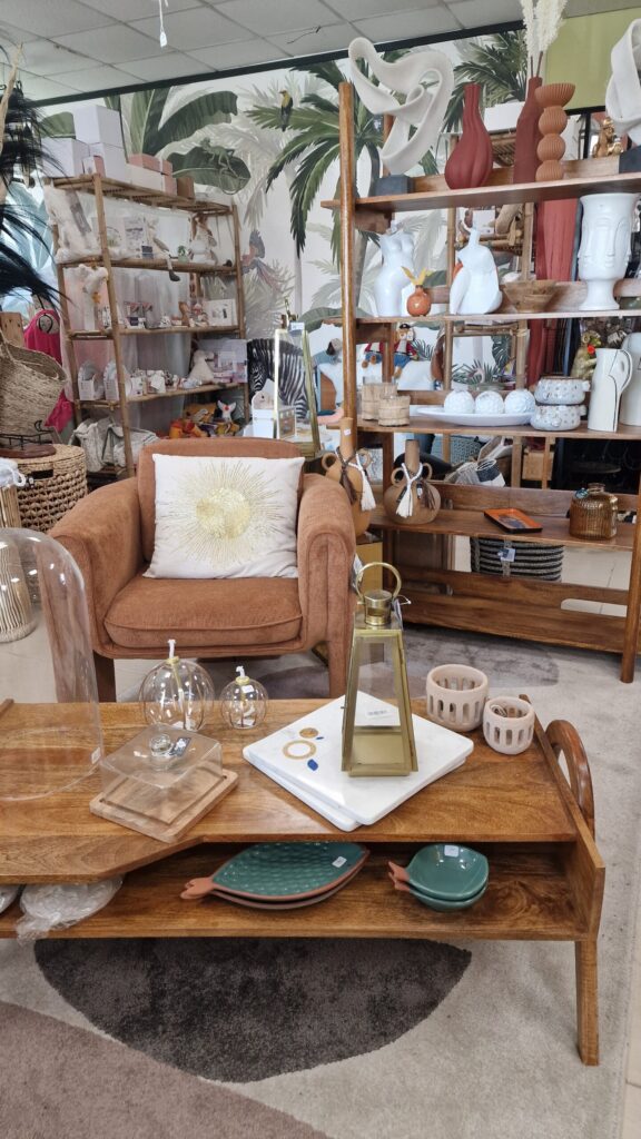 Coin détente dans la boutique Merci Cheri à Auterive, avec un fauteuil confortable, une table basse en bois ornée de décorations en verre et céramique, et des étagères